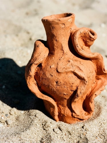 A close-up of a vibrant terracotta sculptural vase with organic curves, resting on a soft sand surface under harsh direct sunlight, casting a sharp dramatic shadow, bold editorial photography, 35mm lens.