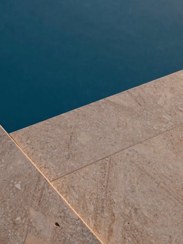 Close-up of premium warm travertine pool coping meeting deep blue water, textured natural stone, soft twilight lighting, architectural photography