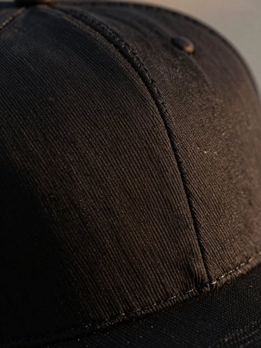 Extreme macro shot of a structured black cap crown, showing the precise, heavy-gauge stitching under warm side lighting, highlighting the texture against deep shadows.