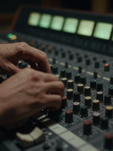 Moody close-up of hands adjusting dials on an analog mixing console in a dimly lit recording studio, soft green ambient light glowing from the level meters, 35mm film grain, high-contrast shadows