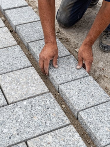 Tight overhead shot of stone paving installation in progress, worker hands placing granite sett blocks into a sand bed, natural daylight, aggregate texture and joint lines sharp, construction site context visible at frame edges