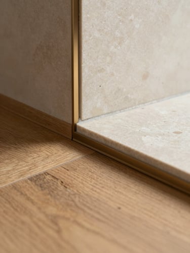 Straight-on macro shot of a precise material transition, oak flooring meeting travertine cream tiles with a perfect brushed bronze joint, soft natural side-light highlighting the matte textures.