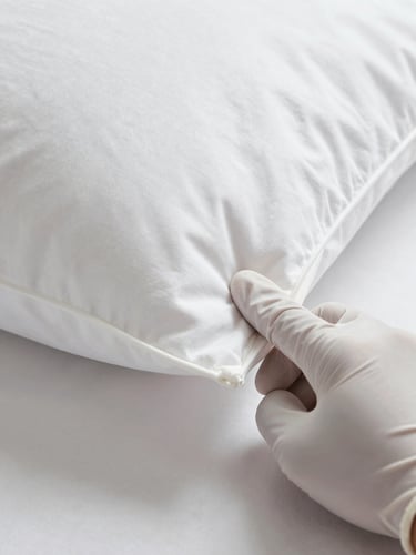 Extreme close-up of a gloved hand pressing a fingertip precisely into the corner seam of a hotel pillow during a quality assessment, controlled studio lighting from above, white linen surface, sharp focus on the contact point, the rest of the frame dissolving into clean white