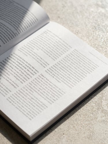 Close-up overhead of a typeset editorial spread laid flat on a pale stone surface, north-facing daylight raking across the pages, ink deep and precise, generous white margins visible