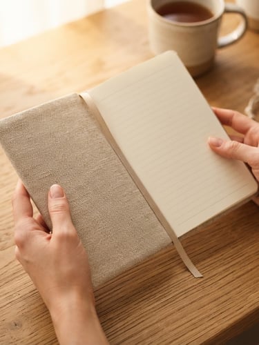 Close-up of hands holding a clean linen-covered planner on an oatmeal-toned wooden desk, soft morning window light, warm shadows, 35mm