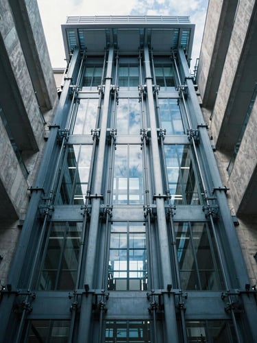 Symmetrical low-angle photograph of a modern glass elevator shaft rising through a concrete and steel atrium, sharp daylight filtering from above