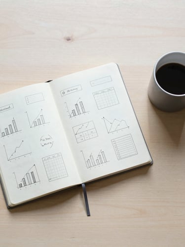 Overhead flat lay of a clean wooden desk with a minimalist notebook containing neat hand-drawn charts, a matte grey coffee mug, soft morning light casting gentle shadows