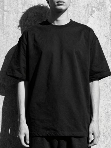 Low-angle high-contrast monochrome fashion shot of a model standing against a raw concrete pillar, wearing a structured heavy-weight oversized black t-shirt, harsh sunlight casting deep geometric shadows