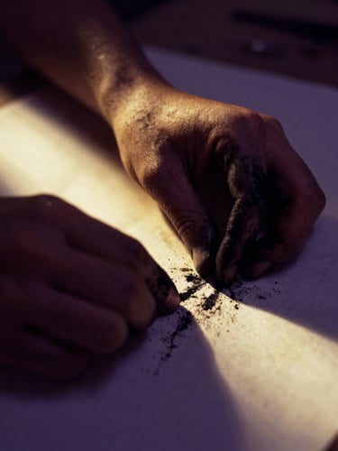 Moody fine-art photography, close-up of hands covered in black charcoal dust working on raw textured paper, dramatic chiaroscuro lighting, warm gold rays cutting through deep violet shadows