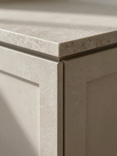 Extreme close-up of a cabinetry joint where a matte stone countertop meets a handleless door panel, overcast natural light raking across the surface to reveal material grain and construction precision—no hands, no props