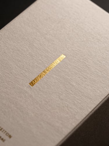 Macro detail of high-end consumer goods packaging, heavy textured paper with minimalist gold foil embossing, dramatic side lighting.