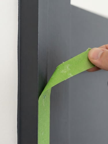 Macro architectural close-up of a razor-sharp paint line against dark charcoal trim, clean green painters tape being peeled back, bright daylight, 35mm lens