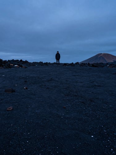 Extreme wide environmental shot of a solitary figure standing on a vast dark volcanic beach at twilight, cool blue hour lighting, deep negative space.