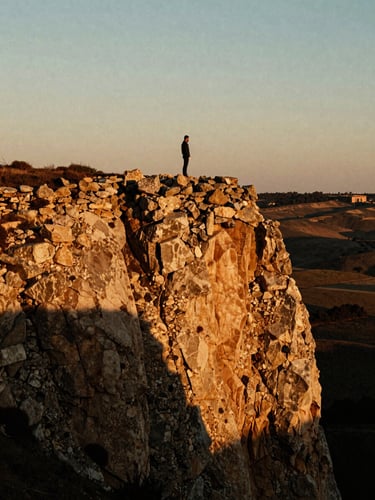 A solitary figure standing on a high rustic stone cliff at golden hour, deep shadows stretching across the landscape, dramatic chiaroscuro lighting, warm amber tones, 35mm film grain