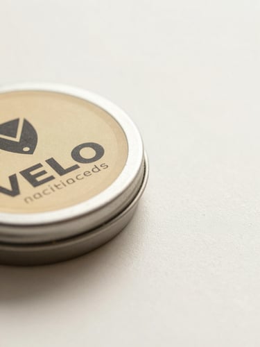 Close-up product flat of VELO nicotine pouch tin, angled at 45 degrees on a clean cream-white surface, studio diffuse lighting with soft rim highlight on the metallic lid edge, minimal negative space on the right