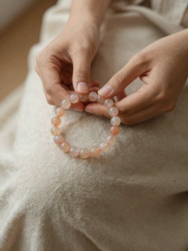 A close-up of a woman's hands resting on her lap, gently holding a soft peach chalcedony bead bracelet, warm diffused morning light, linen texture, shallow depth of field.