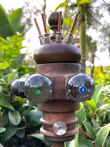 Found object robot head with glass eyes, one green pupil and a larger blue pupil.