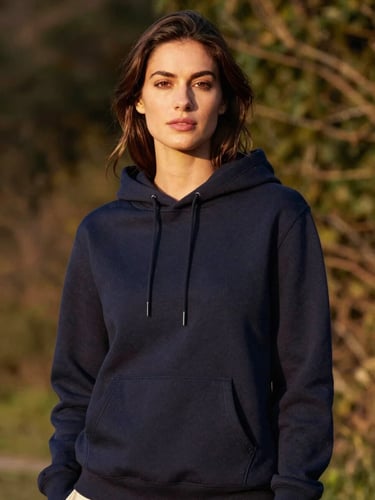 Medium-close portrait of a woman standing confidently in a structured deep navy hoodie, warm golden-hour side light, soft natural foliage background