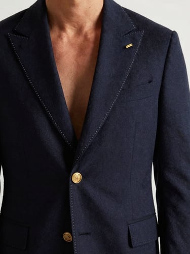 Vertical portrait of a gentleman wearing a perfectly draped bespoke navy blazer, hand-stitched gold lapel buttonhole detail, warm side lighting, clean white background.