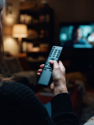 Close-up over-the-shoulder shot of a hand holding a sleek black remote control, soft blue ambient television glow illuminating the buttons, warm domestic interior background, 35mm photography