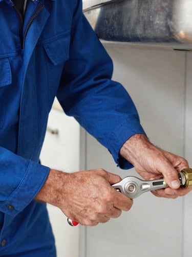 Portrait shot of a professional plumber in a clean blue uniform tightening a copper pipe joint under a kitchen sink, bright natural light, sharp focus on hands and tool.