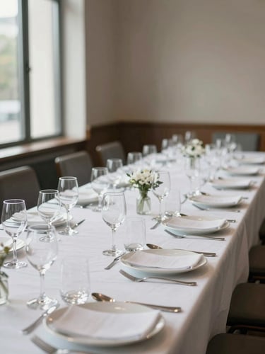 Environmental wide shot of a banquet table fully dressed before service: white linen, arranged glassware and cutlery in precise rows, soft daylight entering from windows on the left, no people, shallow depth of field on the nearest place setting, neutral background of a corporate event hall