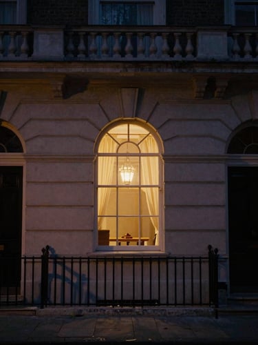 Cinematic editorial photography of a dimly lit classic London townhouse facade at twilight, warm ambient light glowing from a single arched window, deep shadows, 35mm film grain