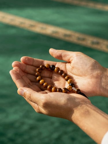 Close-up of clean hands holding a wooden prayer beads inside a sunlit mosque with green carpet, warm editorial photography style.