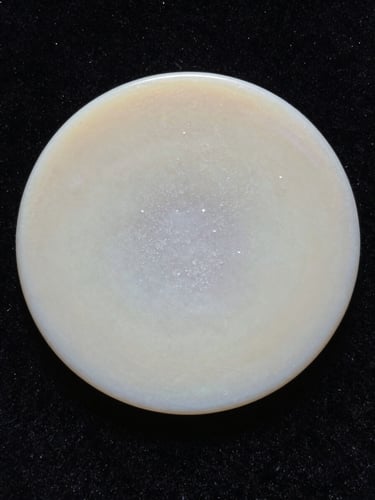 Extreme close-up of a pale wax disc resting on black velvet, soft directional light from the left revealing crystalline surface texture, no labels, no hands, no face — purely material