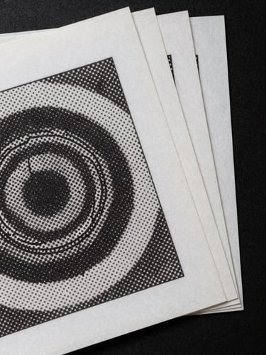 Extreme close-up overhead flat-lay of printed color proof sheets fanned across a matte black surface, raked side-light revealing ink sheen and paper grain, one sheet showing a halftone rosette detail at 1:1 magnification, deep shadow at edges