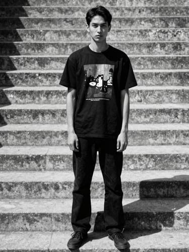 Gritty editorial streetwear portrait of a model standing on brutalist concrete steps, wearing a black graphic t-shirt, harsh direct sunlight, high-contrast shadows