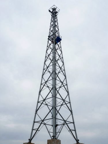 Wide environmental shot from below looking up a three-legged self-support steel lattice cell tower at full height, tower legs anchored in concrete footings at lower frame, overcast daylight sky behind, royal blue safety equipment visible on one leg, no foliage obstruction, cold steel tones dominating