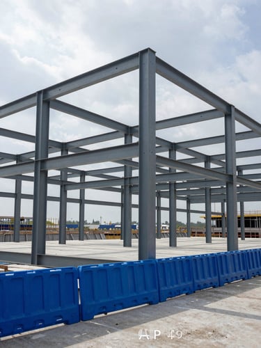 Mid-range environmental shot of a commercial building structural steel frame at mid-construction, royal blue safety barriers along the base, overcast daylight casting even shadows across the exposed beam grid, concrete slab visible at grade level, site context showing scale of the structure