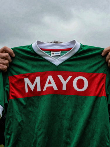 Close-up, weathered hands holding a vintage Mayo GAA jersey, worn fabric, dramatic overcast Irish daylight, high contrast, 35mm film, rich green and red tones.