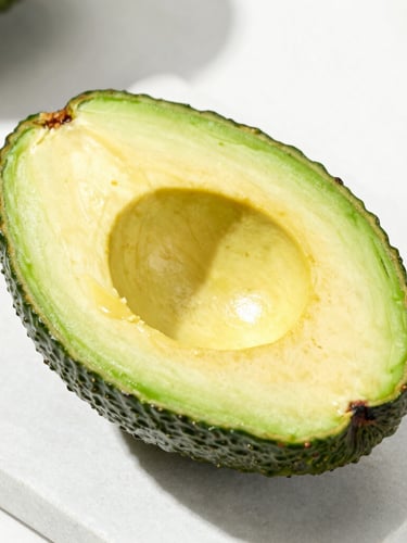 A pristine sliced Hass avocado showing its rich buttery texture and green skin, resting on a clean white stone surface, bright natural daylight, 50mm