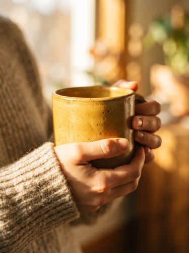 Close-up of a warm yellow stoneware mug held by hands with natural sunlight, bright golden-hour window light, tactile glaze textures