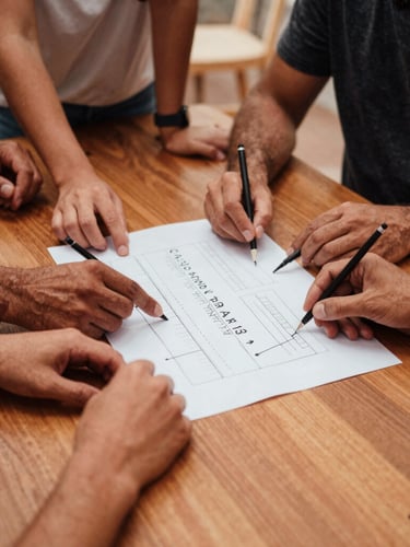 Wide environmental shot of a collaborative workshop around a wooden table, hands mapping out a strategy on paper, warm terracotta tones, natural daylight