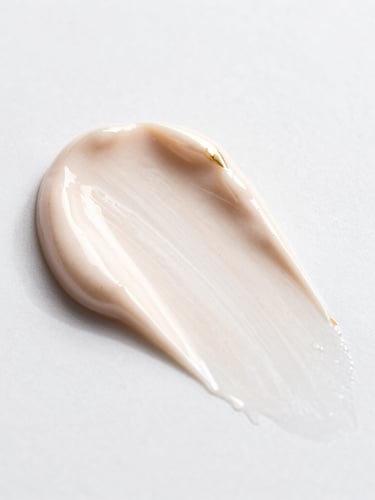 Macro texture shot of rich cosmetic cream swatched on a clean white surface, soft directional studio lighting, champagne gold accents