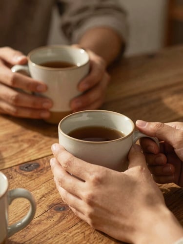 Close-up of hands holding warm ceramic mugs around a rustic cedar table, soft natural golden hour light.