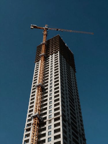 Low-angle heroic photograph of a modern high-rise tower under construction, towering cranes against a deep blue sky, sharp architectural daylight