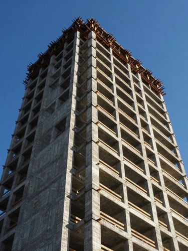 Low-angle architectural photograph of a modern high-rise concrete tower frame under construction against a clear blue sky, sharp daylight shadows.