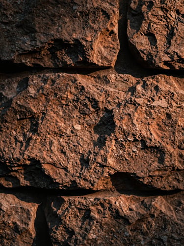 Extreme close-up of a rough stone wall surface, warm raking side-light casting deep shadows across the texture, terracotta and charcoal tones, no people, tactile and still, art-film editorial quality
