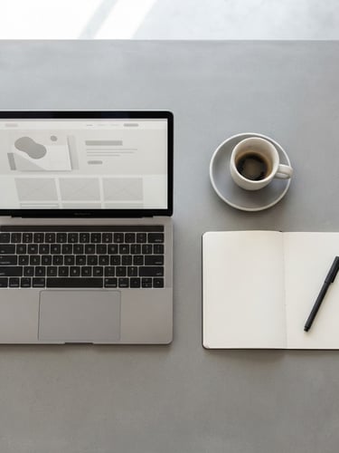 A minimalist overhead flat lay of a clean workspace with a laptop displaying a modern website mockup, a notebook, and a ceramic coffee cup on a neutral grey table, bright soft studio light.