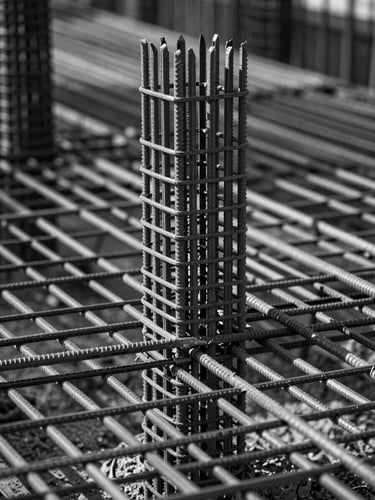 Sharp, straight-on architectural photograph of a reinforced steel rebar grid before a concrete pour, dramatic raking side-light highlighting the metallic geometry, desaturated slate tones.