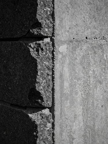 Extreme close-up of a structural masonry joint where hand-chiseled dark stone meets smooth gray board-formed concrete, perfect alignment, high-contrast side-light.
