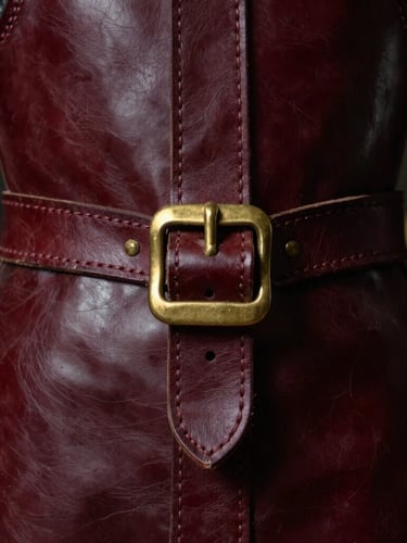 Macro shot of thick hand-stitched leather seams on deep burgundy armor, heavy brass buckles, atmospheric low-key workshop lighting