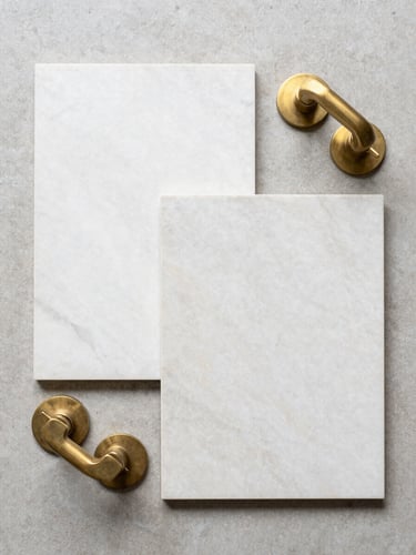 Top-down flat lay of honed marble tile samples and solid-brass fixtures on a neutral concrete background, bright diffused daylight, sharp focus