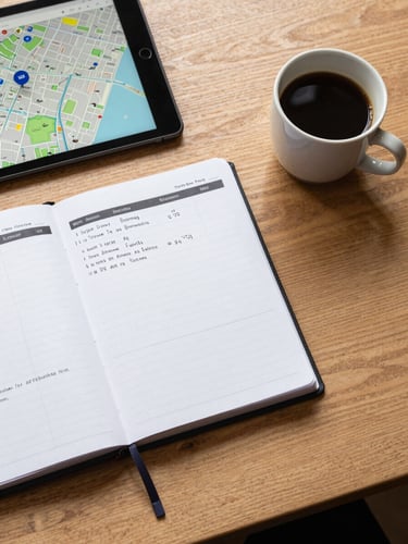 A top-down view of a clean wooden desk in London, containing an organized planner, a tablet showing a local map with a pinned business, and a ceramic cup of coffee, bright overcast daylight