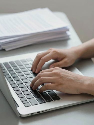 Close-up of hands typing on a laptop keyboard alongside neatly stacked paper documents on a slate grey desk, neutral bright daylight