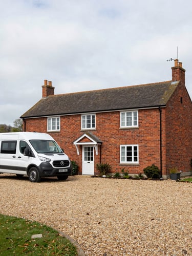 A wide environmental shot of a clean, simple brick guesthouse in Bedford with a large gravel driveway where a commercial transit van is parked, flat overcast daylight.